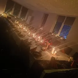 Długi stół nakryty białym obrusem z dekoracjami świątecznymi, świecami i czerwonymi serwetkami w kształcie stożków, oświetlony ciepłym światłem z dekoracyjnej lampki na suficie, widok na nocne...