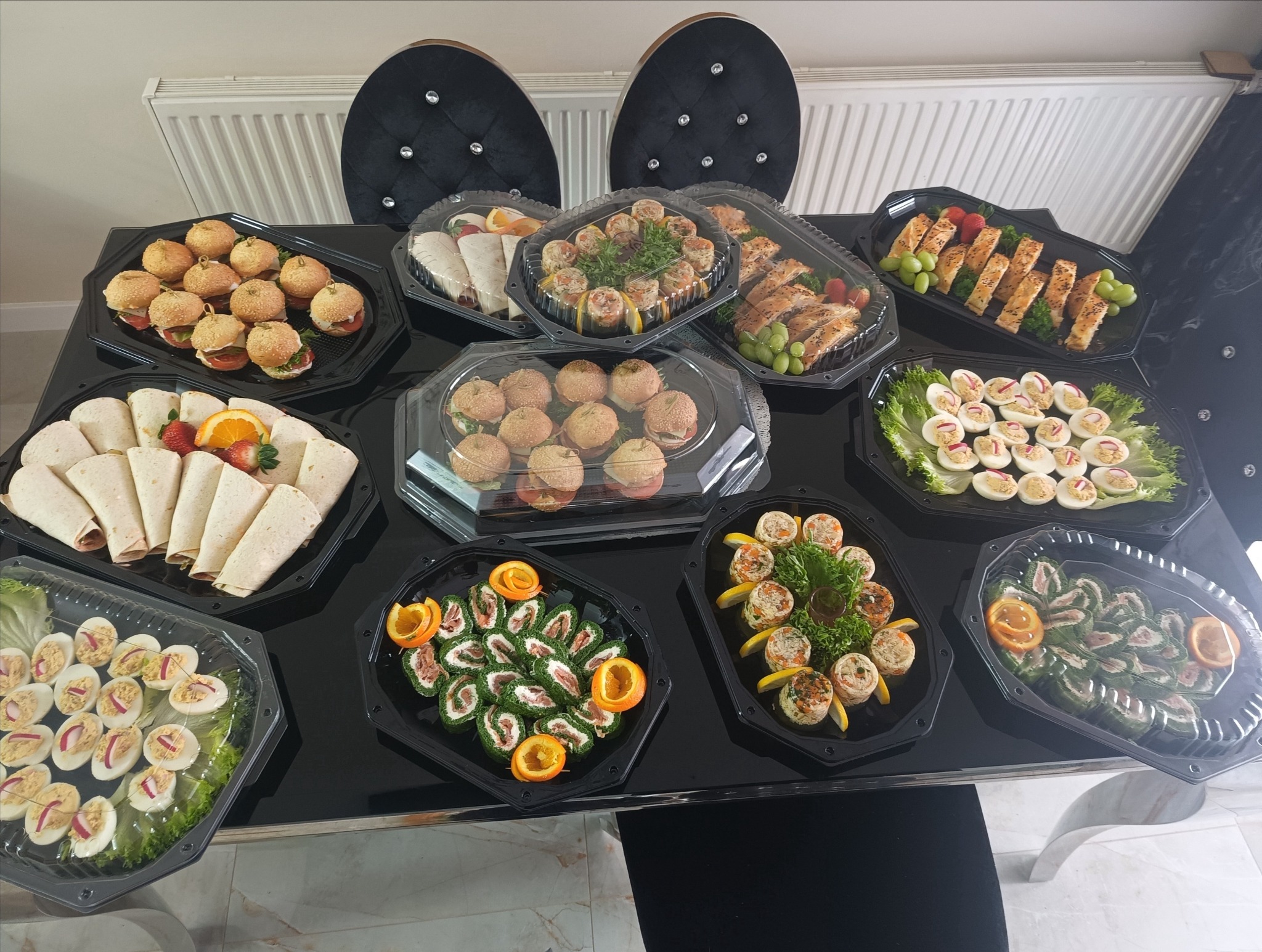 Ustawienie stołu z różnorodnymi przekąskami cateringowymi w czarnych, plastikowych pojemnikach: mini burgery, roladki z tortilli, jajka faszerowane, roladki z łososiem i szpinakiem, oraz ciasto...