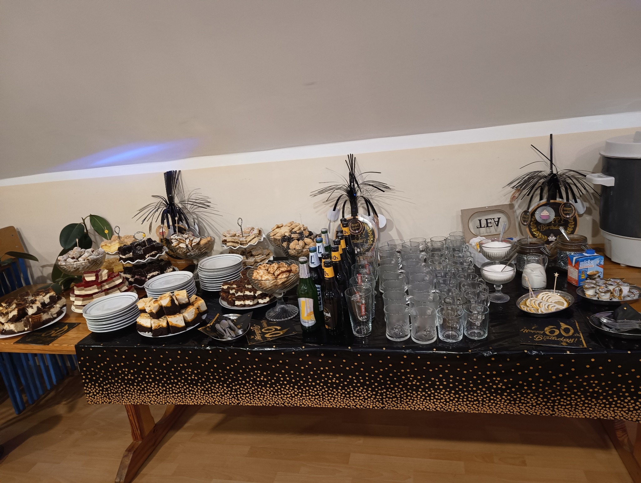 Stół z cateringiem na 60. urodziny: różnorodne ciasta, ciasteczka, napoje w szklanych naczyniach, dekoracje urodzinowe w czarno-złotej kolorystyce.