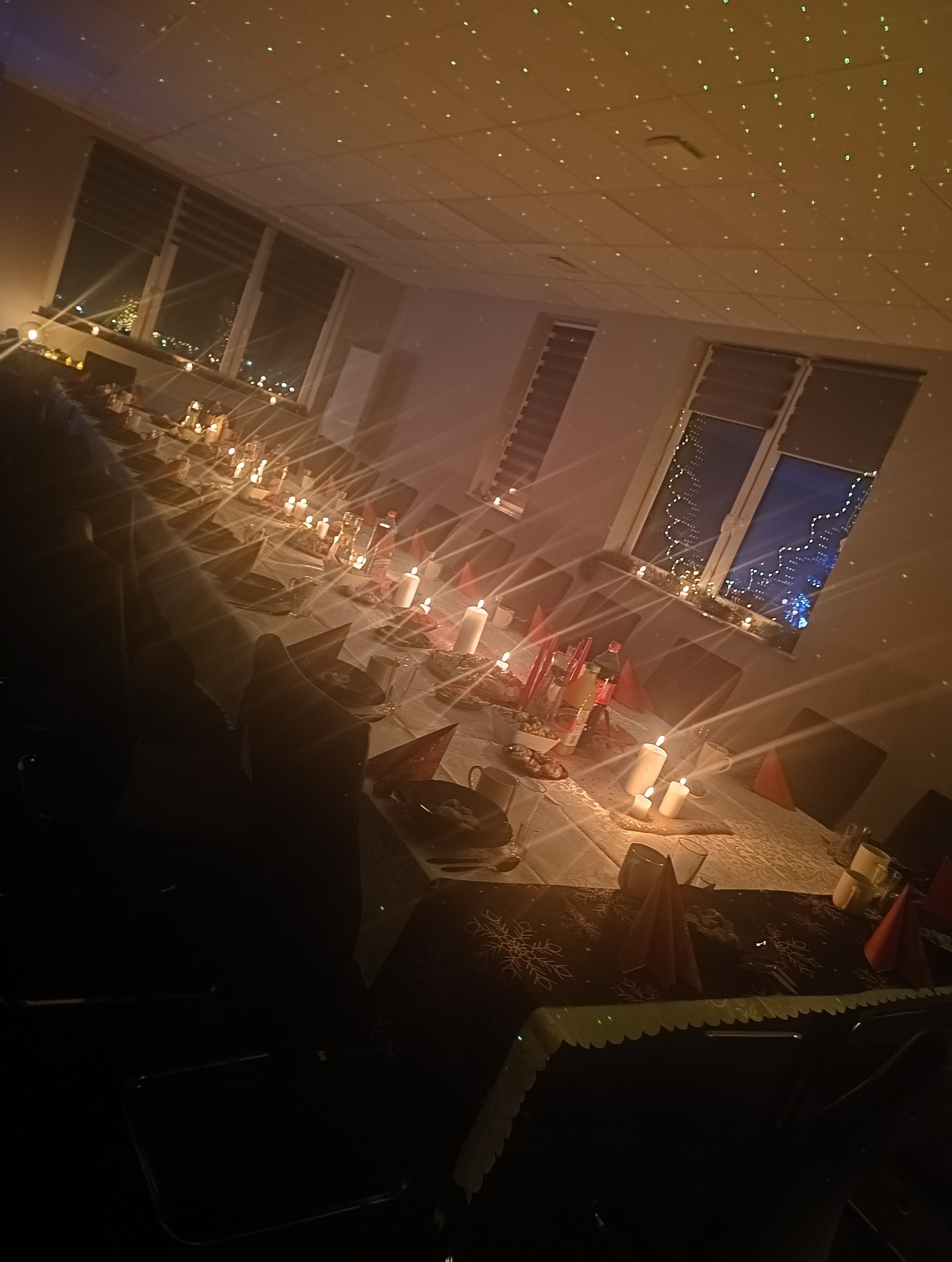 Długi stół nakryty białym obrusem z dekoracjami świątecznymi, świecami i czerwonymi serwetkami w kształcie stożków, oświetlony ciepłym światłem z dekoracyjnej lampki na suficie, widok na nocne...