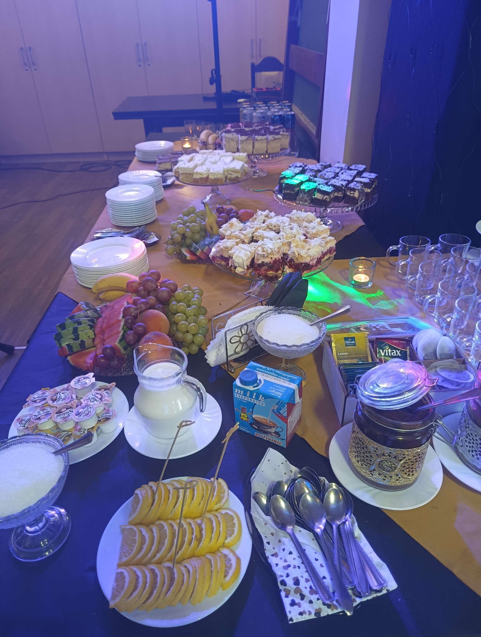 Stół z przygotowanym bufetem: ciasta na paterach, talerze z owocami (winogrona, banany, arbuz, brzoskwinie), dzbanek z mlekiem, miseczki z cukrem, plasterki cytryny na szpilkach, herbata, dżem...