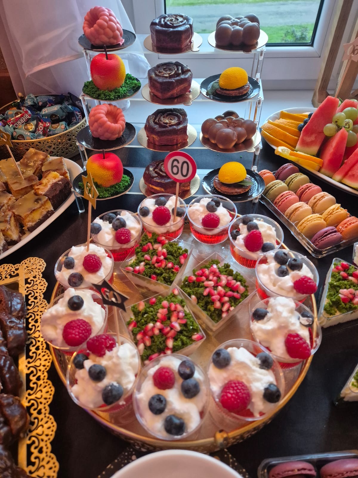 Bogato zastawiony stół z deserami: mini torciki, musy owocowe z borówkami i malinami, makaroniki, owoce. Dekoracja z numerem 60. Elegancki catering na przyjęcie.