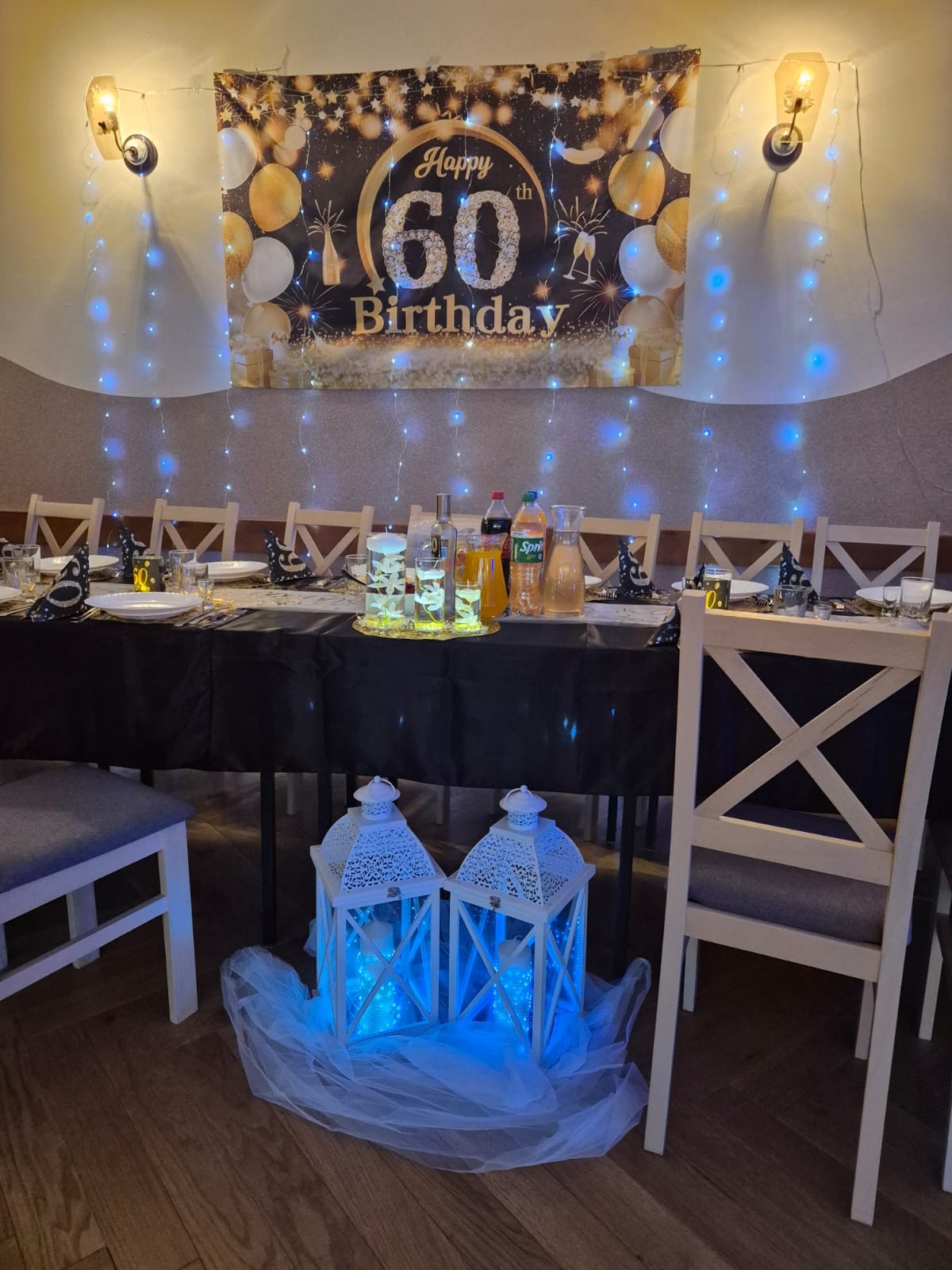 Uroczysty stół na 60. urodziny z dekoracjami: baner, lampki, napoje w dzbankach i lampiony pod stołem, tworzące ciepły, imprezowy klimat.