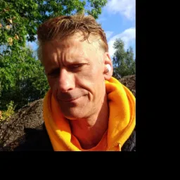 Mężczyzna w żółtej bluzie z kapturem, z bezprzewodową słuchawką w uchu, na tle zieleni i błękitnego nieba.