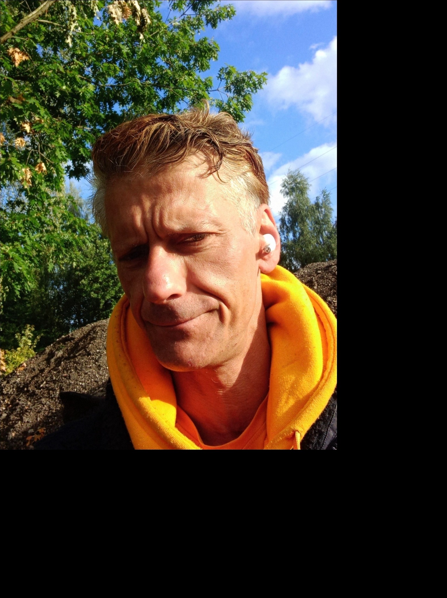 Mężczyzna w żółtej bluzie z kapturem, z bezprzewodową słuchawką w uchu, na tle zieleni i błękitnego nieba.