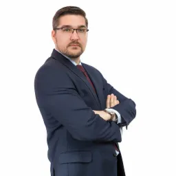 Portret mężczyzny w garniturze z założonymi rękami, na białym tle, z widocznym zegarkiem na lewym nadgarstku.