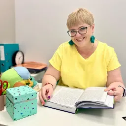 Uśmiechnięta kobieta w okularach, żółtej bluzce i zielonych kolczykach siedzi za biurkiem z książką, obok kolorowe kredki, nożyczki, pudełko i model czaszki z zaznaczonymi kolorami.