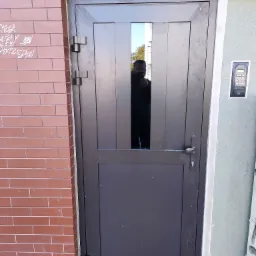 Ciemna, stalowa konstrukcja drzwi wejściowych z szybą, zamontowana w murze z cegły i szarego tynku, widoczne ślady po montażu i graffiti na cegłach.