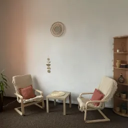 Jasne, minimalistyczne wnętrze gabinetu z dwoma drewnianymi fotelami z poduszkami w kolorze terakoty, małym stolikiem i rośliną doniczkową przy oknie.