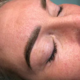 Zbliżenie na twarz kobiety z zamkniętym okiem i świeżo wykonaną stylizacją brwi metodą pudrową, widoczne drobne zaczerwienienia skóry.