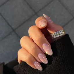 Dłoń z owalnymi paznokciami w kolorze nude, ozdobionymi drobnymi, srebrnymi cyrkoniami, nosząca złoty pierścionek z rzędami cyrkonii, na tle szarej nawierzchni i czarnego materiału ubrania.