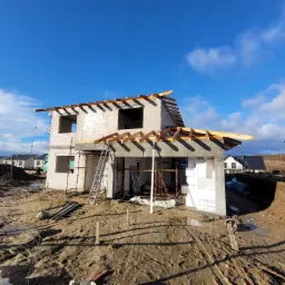 Niedokończony dom jednorodzinny z betonu komórkowego w stanie surowym otwartym, widoczne rusztowania, elementy drewniane więźby dachowej, ocieplenie styropianem, teren budowy z ziemią i materiałami...