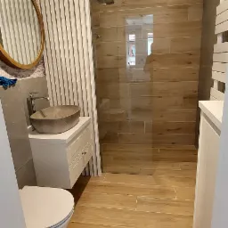 Nowoczesna łazienka z drewnopodobnymi płytkami, kabiną prysznicową typu walk-in, okrągłym lustrem w złotej ramie i umywalką w kształcie kamienia.