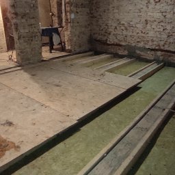 Usługi PSSN - Montaż podłogi na legarach z izolacją w pomieszczeniu z ceglanymi ścianami. Częściowo ułożone płyty OSB. Widoczne narzędzia w tle.