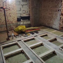 Usługi PSSN - Drewniana konstrukcja pod podłogę z izolacją termiczną w surowym wnętrzu z ceglanymi ścianami. Widoczne narzędzia i lampa robocza.