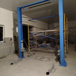 Usługi PSSN - Niebieski podnośnik samochodowy dwukolumnowy w surowym wnętrzu warsztatu, z rusztowaniem w tle. Widoczne okablowanie i elementy instalacji elektrycznej na ścianie.
