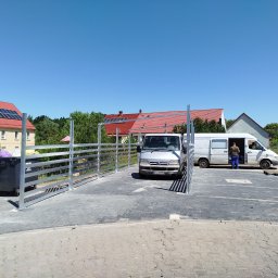 Usługi PSSN - Metalowa konstrukcja ogrodzenia na kostce brukowej, z zaparkowanymi samochodami dostawczymi w tle. Widoczne budynki z czerwonymi dachami i panele słoneczne.