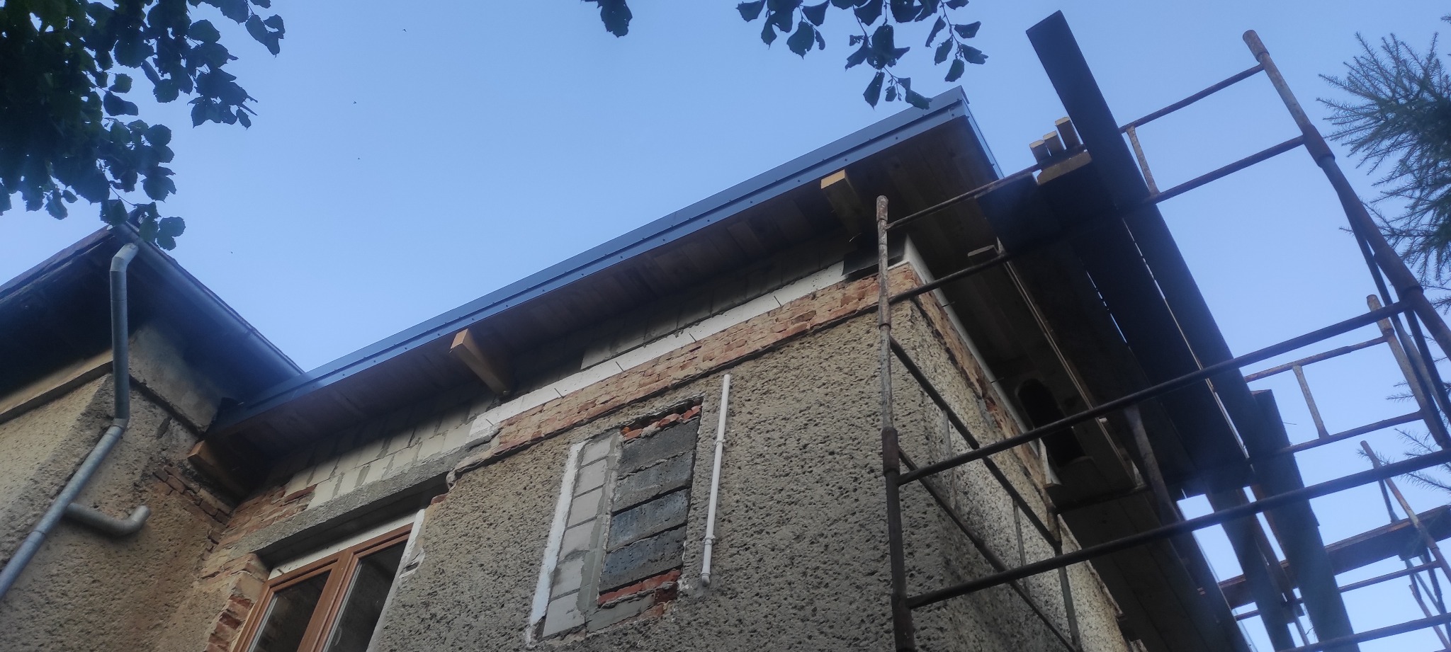 Fragment elewacji domu w trakcie remontu z widocznym rusztowaniem, nową rynną i oknem w trakcie wymiany. Widok z dołu, niebo w tle.