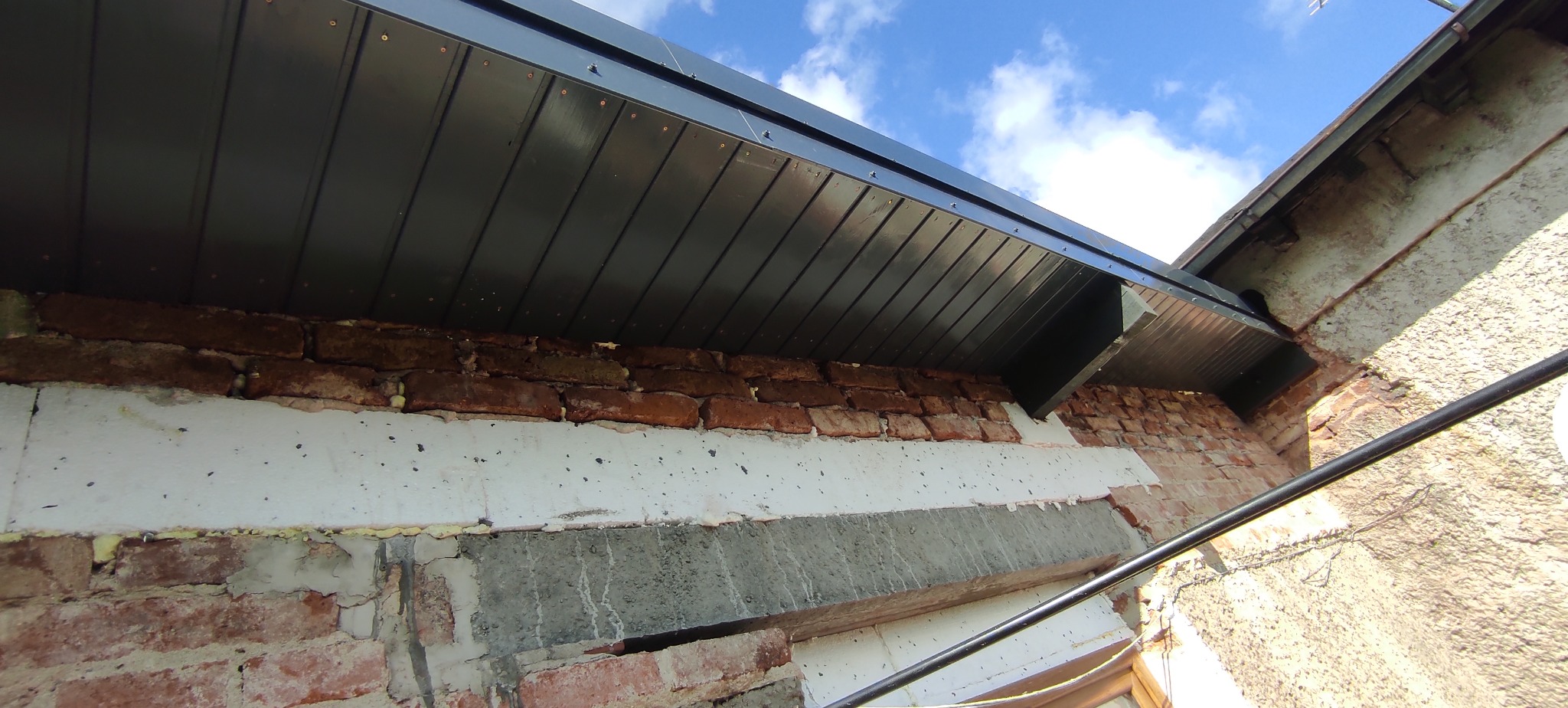 Zamontowana, czarna podbitka dachowa z widocznymi śrubami, na tle fragmentu ściany z cegły i ocieplenia. Ujęcie z dołu, widoczne niebieskie niebo.