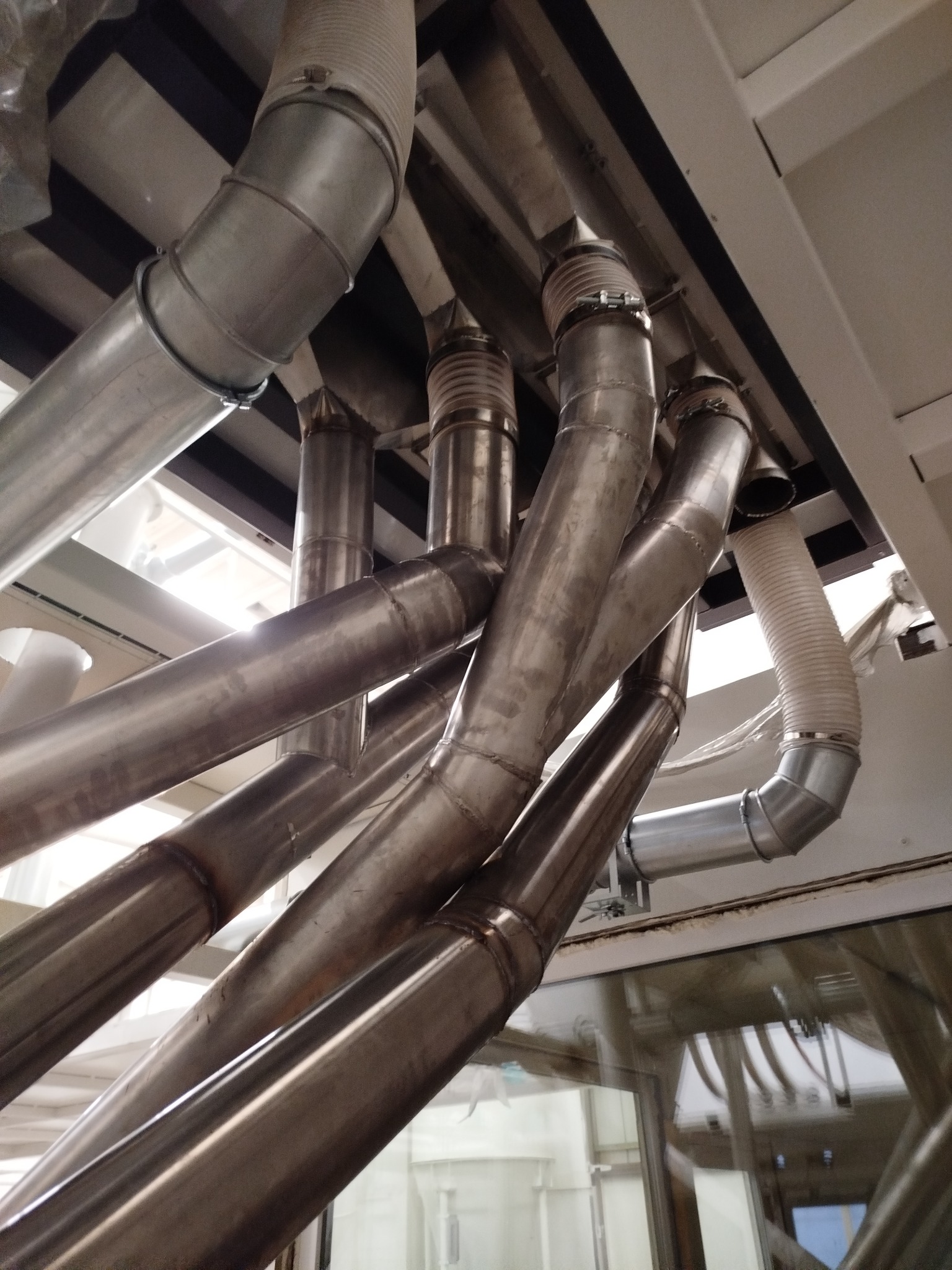 Metalowe rury wentylacyjne z widocznymi spawami i złączkami, zamontowane pod sufitem w hali przemysłowej. Ujęcie z dołu, widoczne elementy konstrukcji budynku.