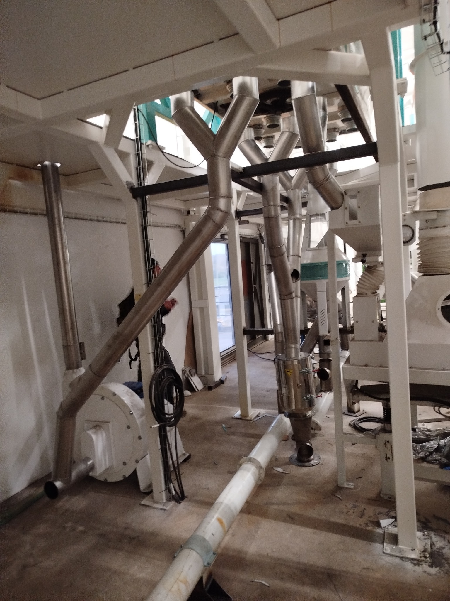 Kompleksowa instalacja przemysłowa z rurami ze stali nierdzewnej, białymi elementami konstrukcyjnymi i widocznymi przewodami, w jasnym, przestronnym wnętrzu hali.