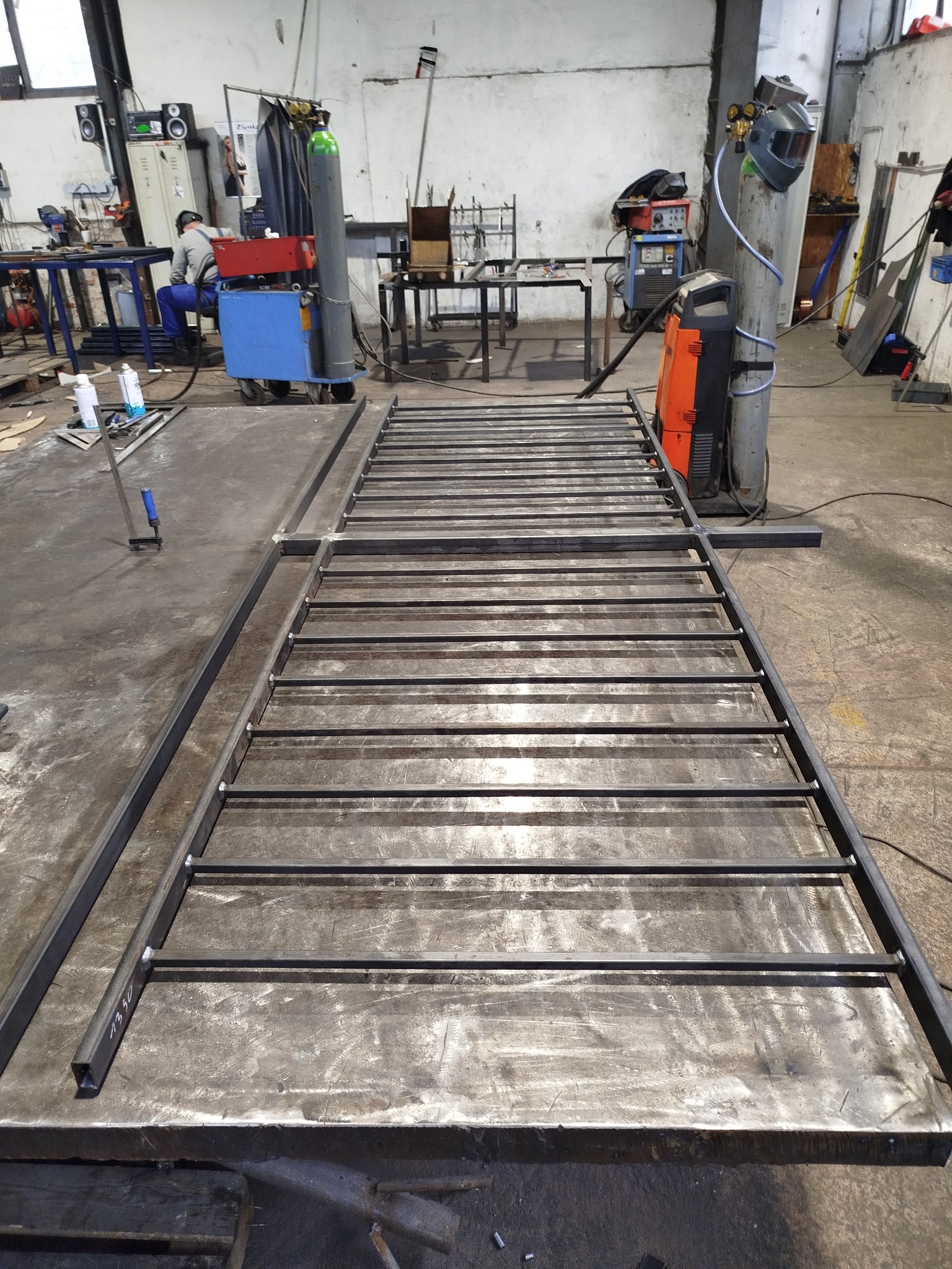 Metalowa konstrukcja spawana z profili, widoczna w warsztacie. Spawarka i butla z gazem w tle. Konstrukcja leży na stole spawalniczym. Widoczne spawy.