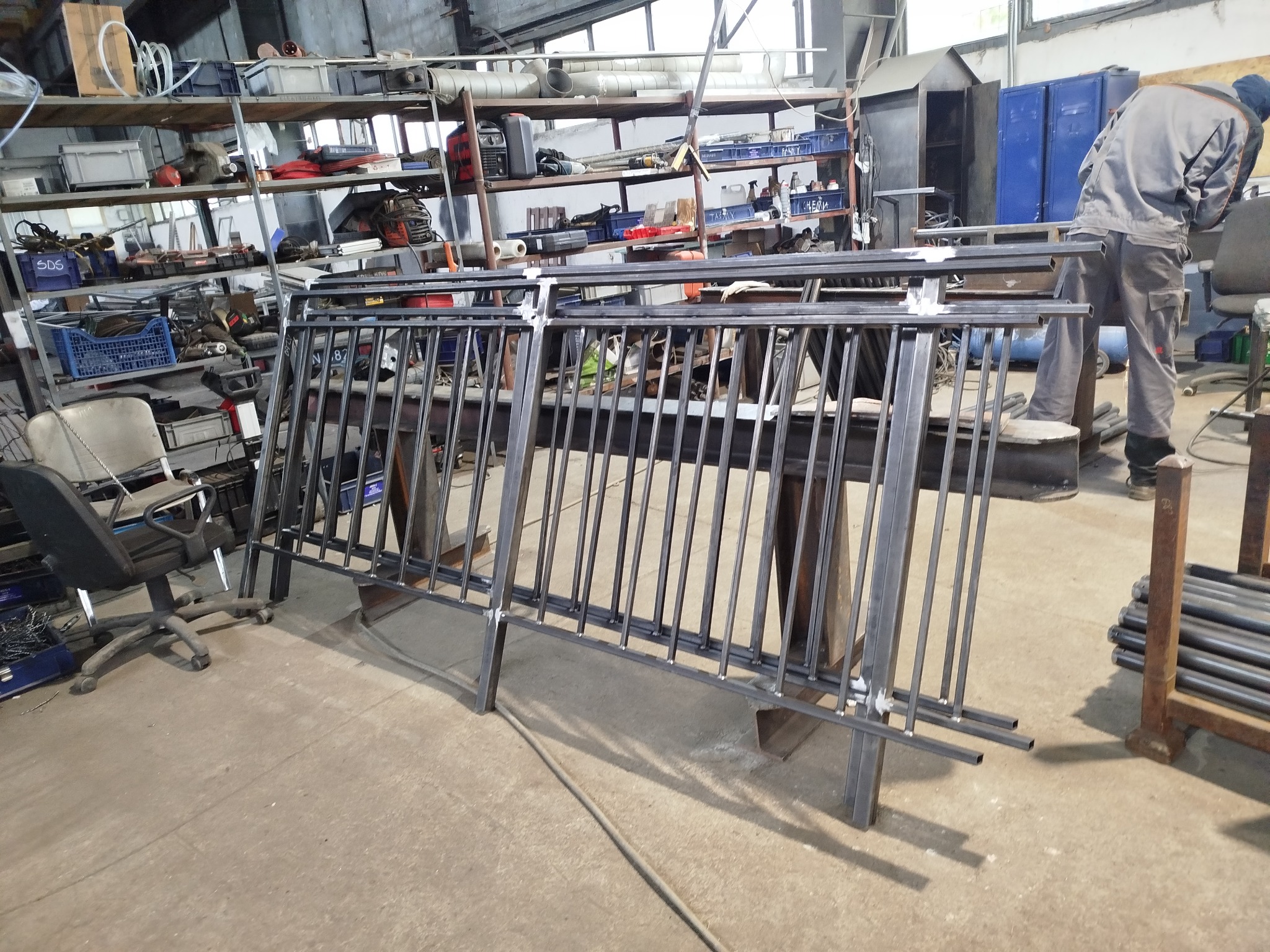 Spawane, surowe, metalowe balustrady w warsztacie, widoczne spawy. W tle półki z narzędziami i spawacz przy pracy. Surowy, industrialny charakter.