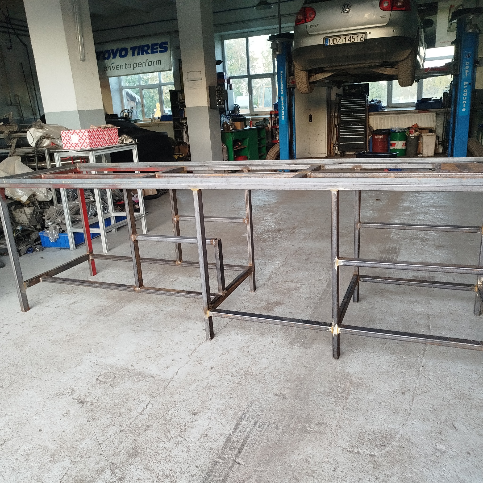 Metalowa konstrukcja spawana w warsztacie, widoczny samochód na podnośniku. Konstrukcja z profili stalowych, surowy stan, widoczne spawy. Warsztat samochodowy w tle.