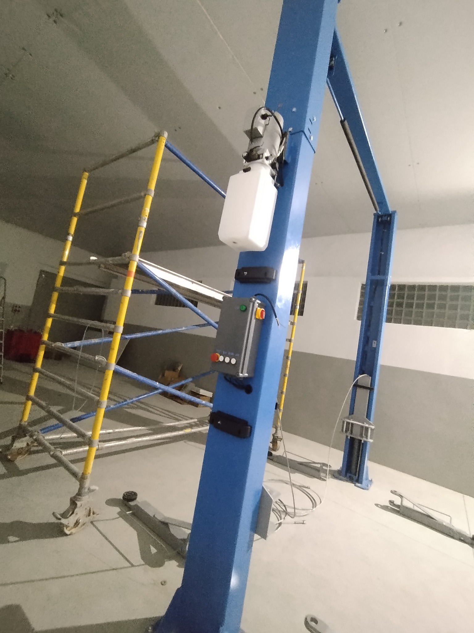 Niebieski podnośnik samochodowy z panelem sterowania w jasnym pomieszczeniu, widoczne rusztowanie i betonowa podłoga. Ujęcie z dołu, oświetlenie sufitowe.