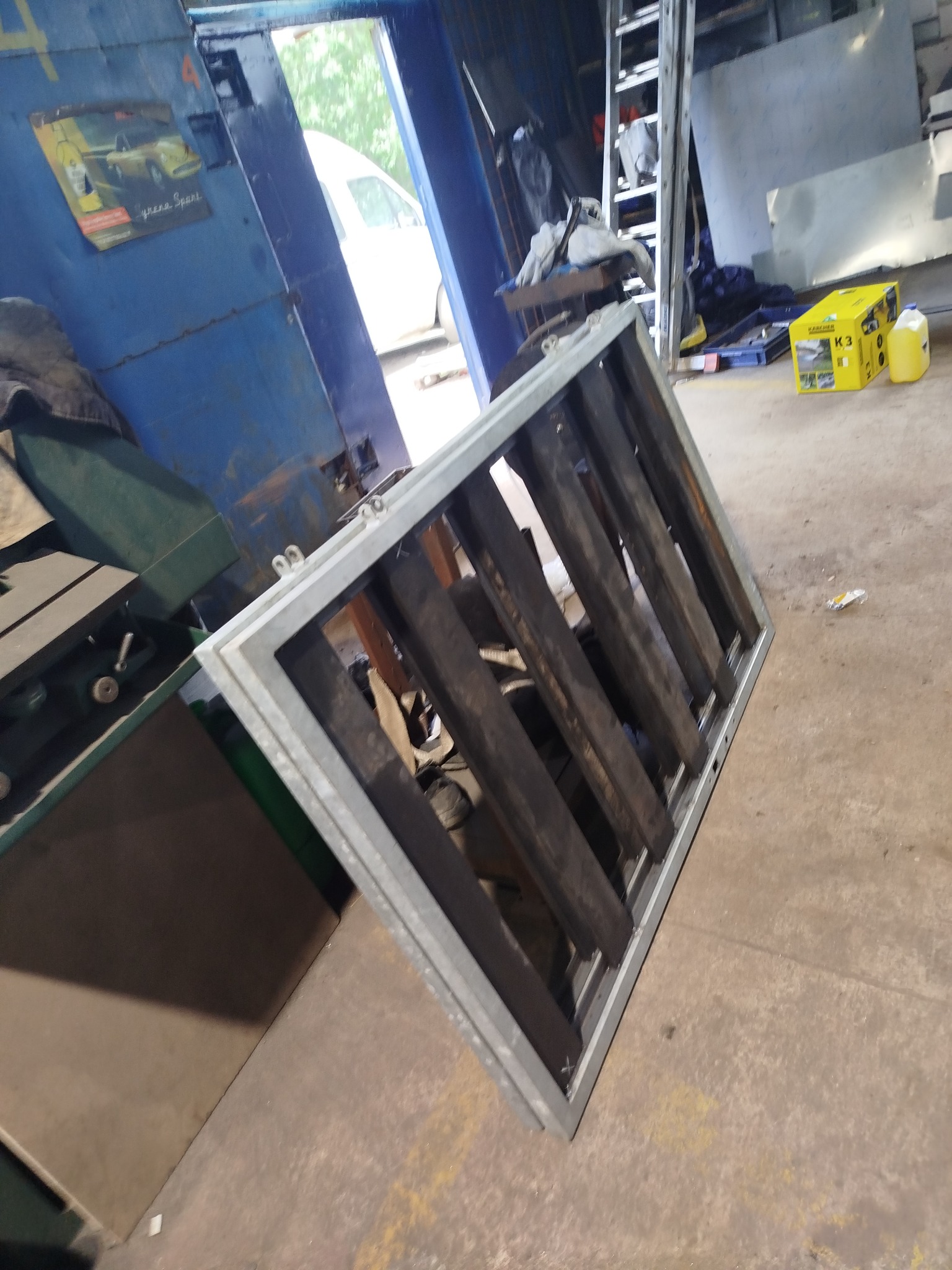 Metalowa konstrukcja kratowa z profili, spawana, w warsztacie. Widoczne narzędzia, drabina i materiały. Niebieska ściana z plakatem w tle.