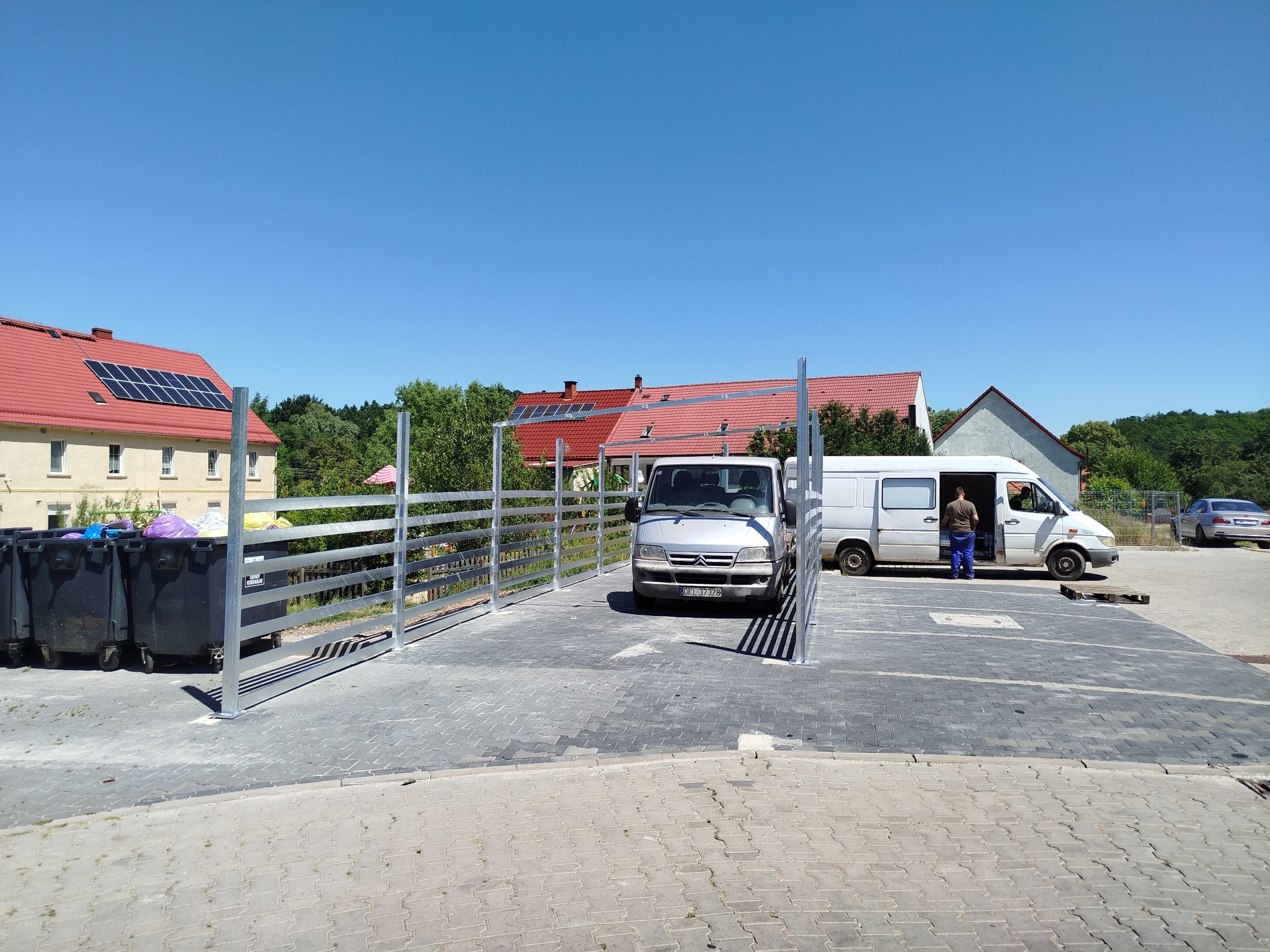 Metalowa konstrukcja ogrodzenia na kostce brukowej, z zaparkowanymi samochodami dostawczymi w tle. Widoczne budynki z czerwonymi dachami i panele słoneczne.