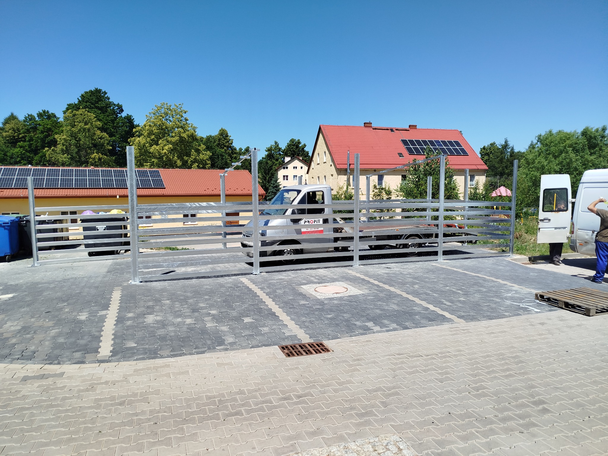 Nowa, metalowa konstrukcja ogrodzenia na posesji z kostki brukowej, z widocznym samochodem dostawczym i budynkami w tle, w słoneczny dzień.