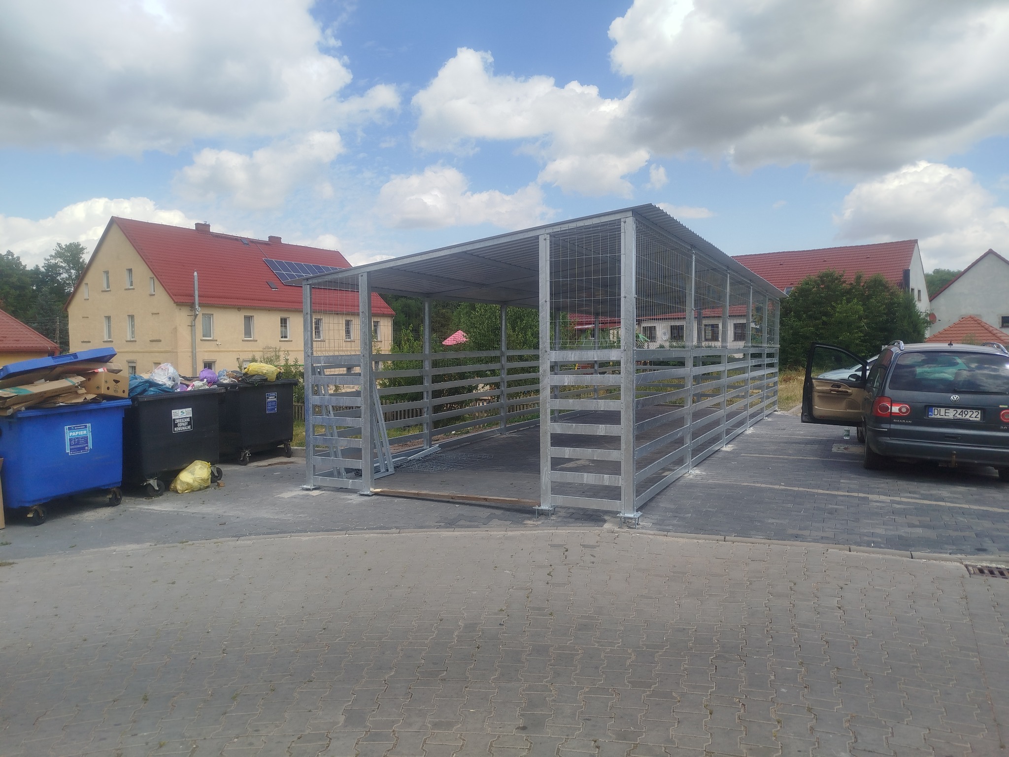 Metalowa konstrukcja w formie wiaty, prawdopodobnie na śmietniki, wykonana ze stalowych profili i siatki, na tle budynków mieszkalnych i błękitnego nieba.