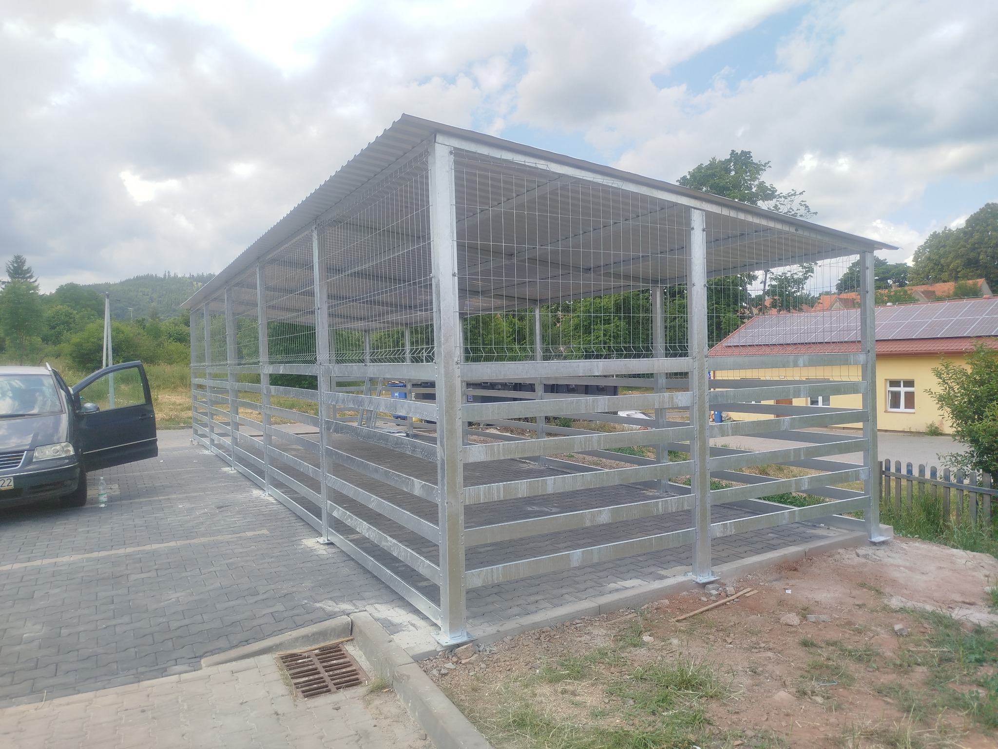 Metalowa konstrukcja zadaszona blachą falistą, prawdopodobnie wiata, na kostce brukowej. W tle budynek z panelami słonecznymi i otwarty samochód osobowy.