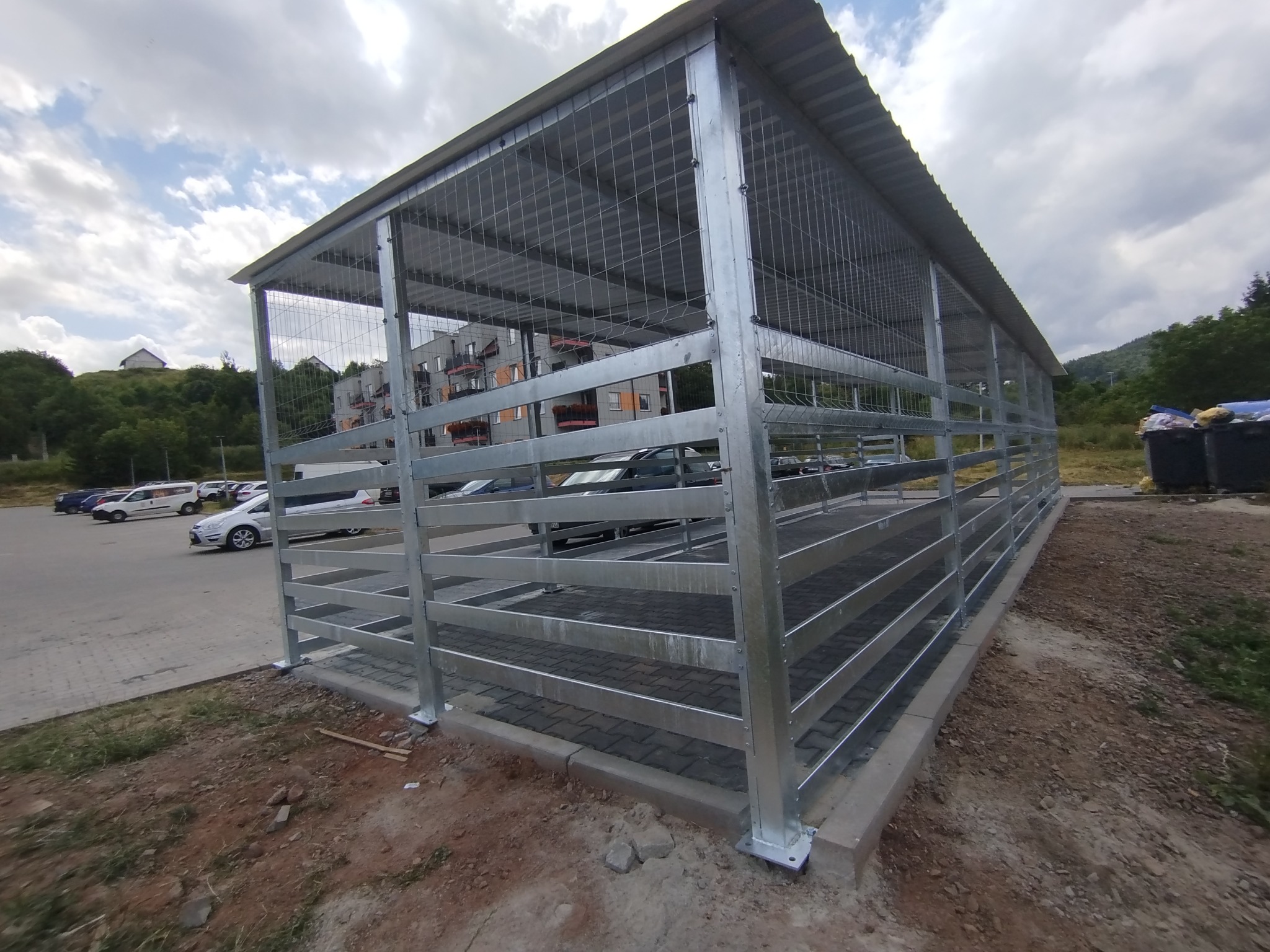 Metalowa konstrukcja z siatką i zadaszeniem, prawdopodobnie wiata śmietnikowa, na parkingu z budynkami w tle, wykonana ze stali ocynkowanej.