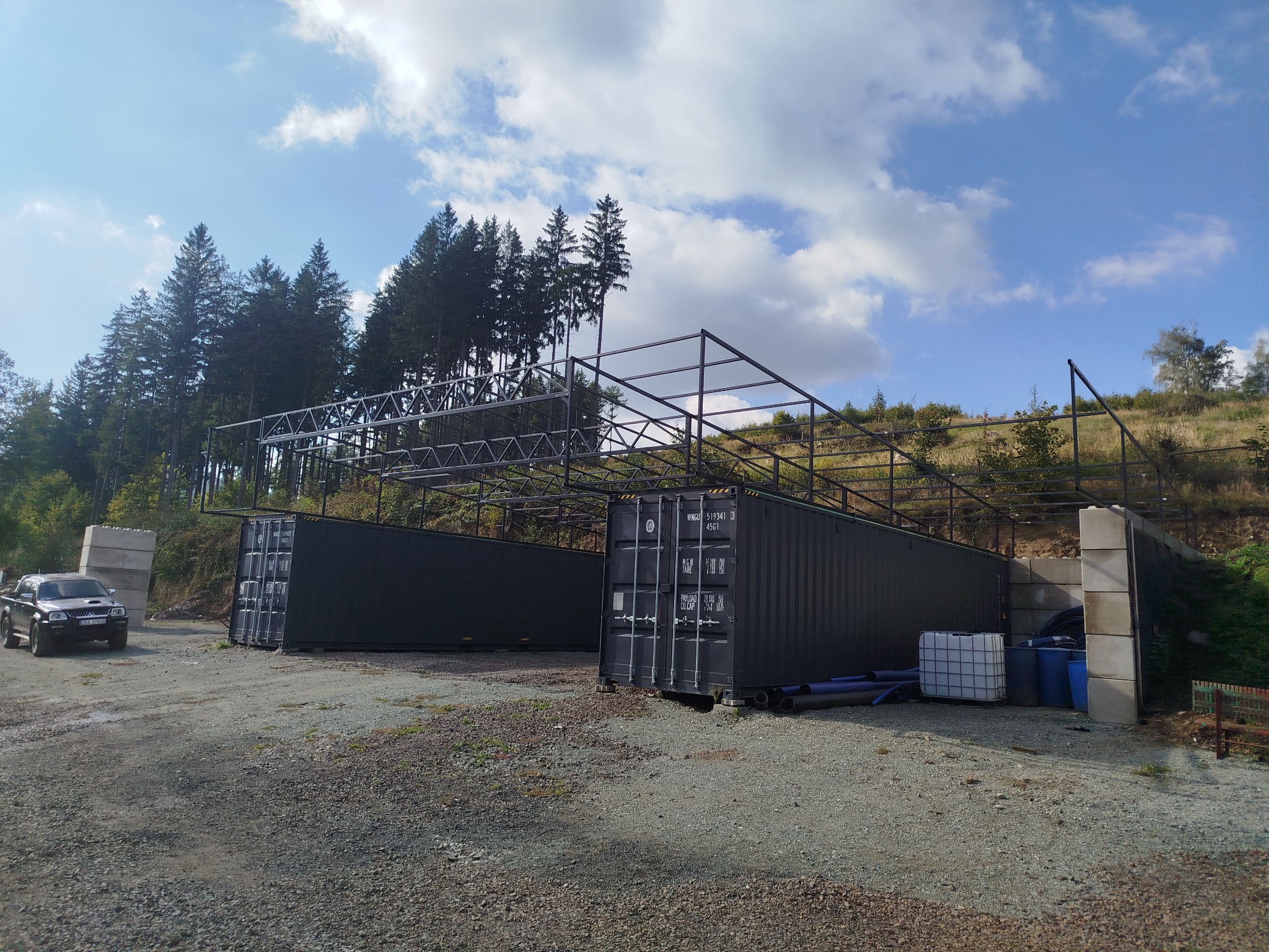 Dwa czarne kontenery morskie z metalową konstrukcją na zewnątrz, otoczone drzewami i gruzem. Widoczny samochód terenowy i białe zbiorniki na wodę.