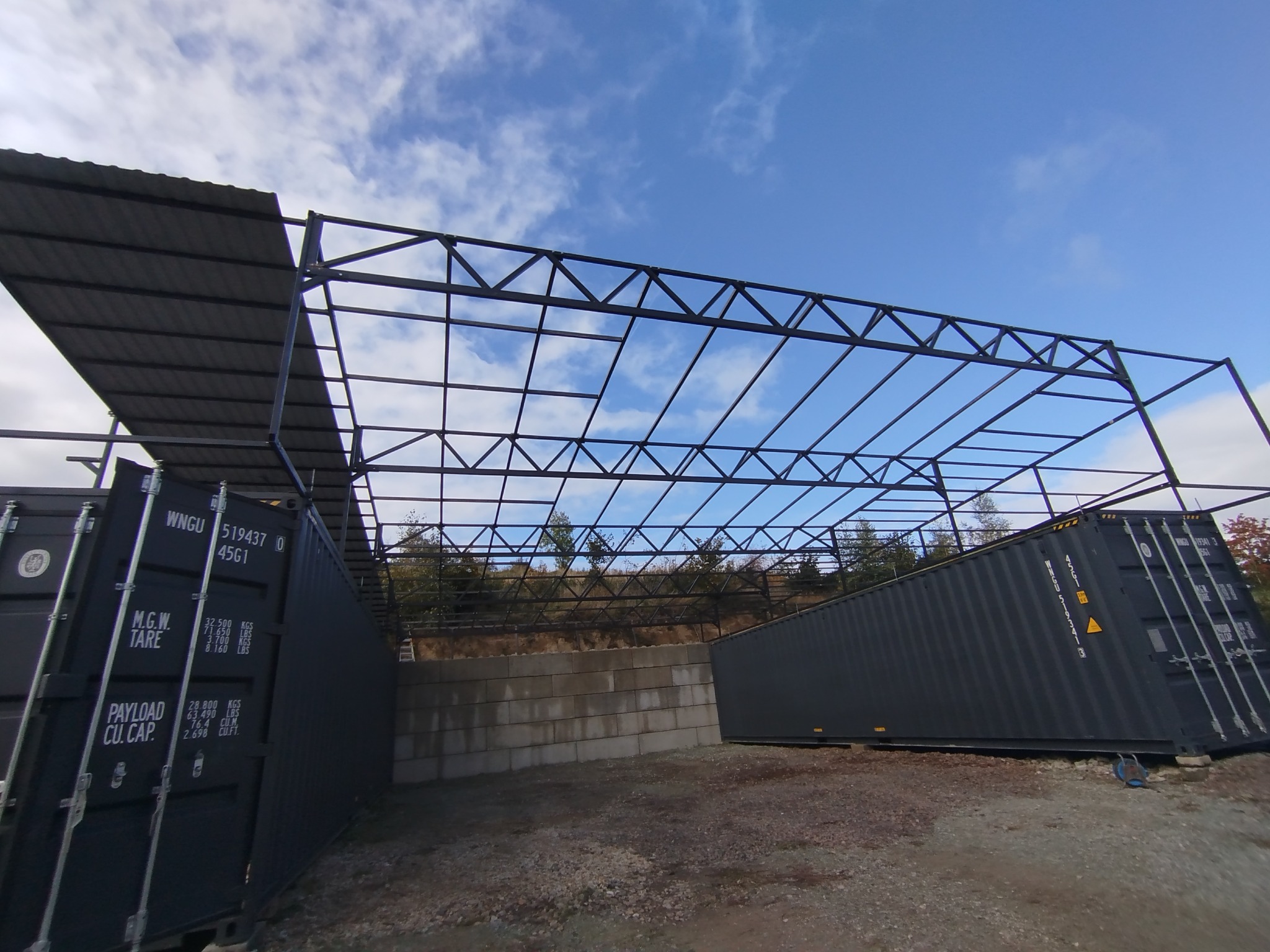Konstrukcja stalowa zadaszenia nad kontenerami transportowymi na tle błękitnego nieba. Widoczne elementy spawane i śruby mocujące. Nietypowe wykorzystanie kontenerów.