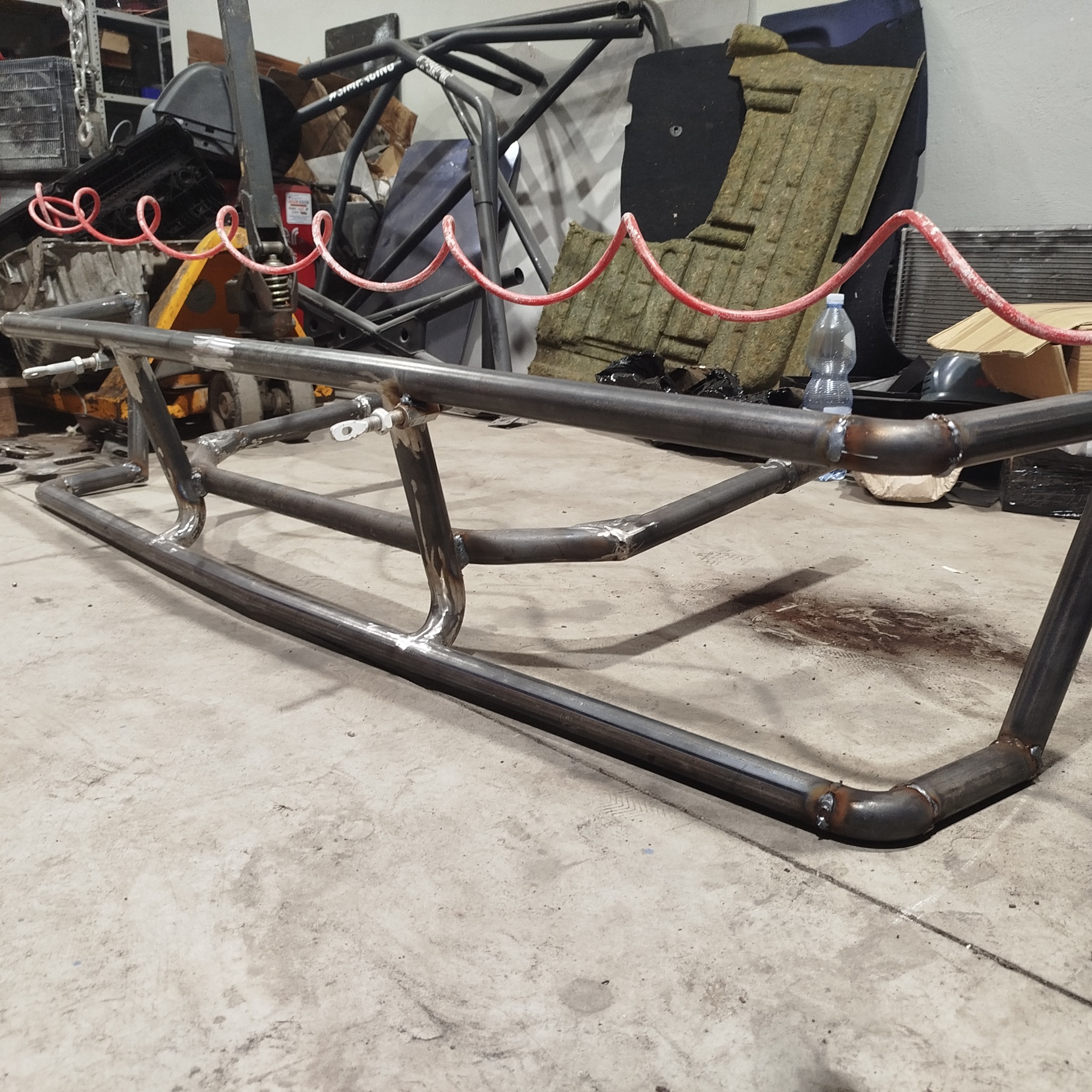 Spawana konstrukcja z rur stalowych na betonowej posadzce, widoczne spawy. W tle narzędzia i materiały warsztatowe, czerwony przewód pneumatyczny. Surowy, industrialny klimat.