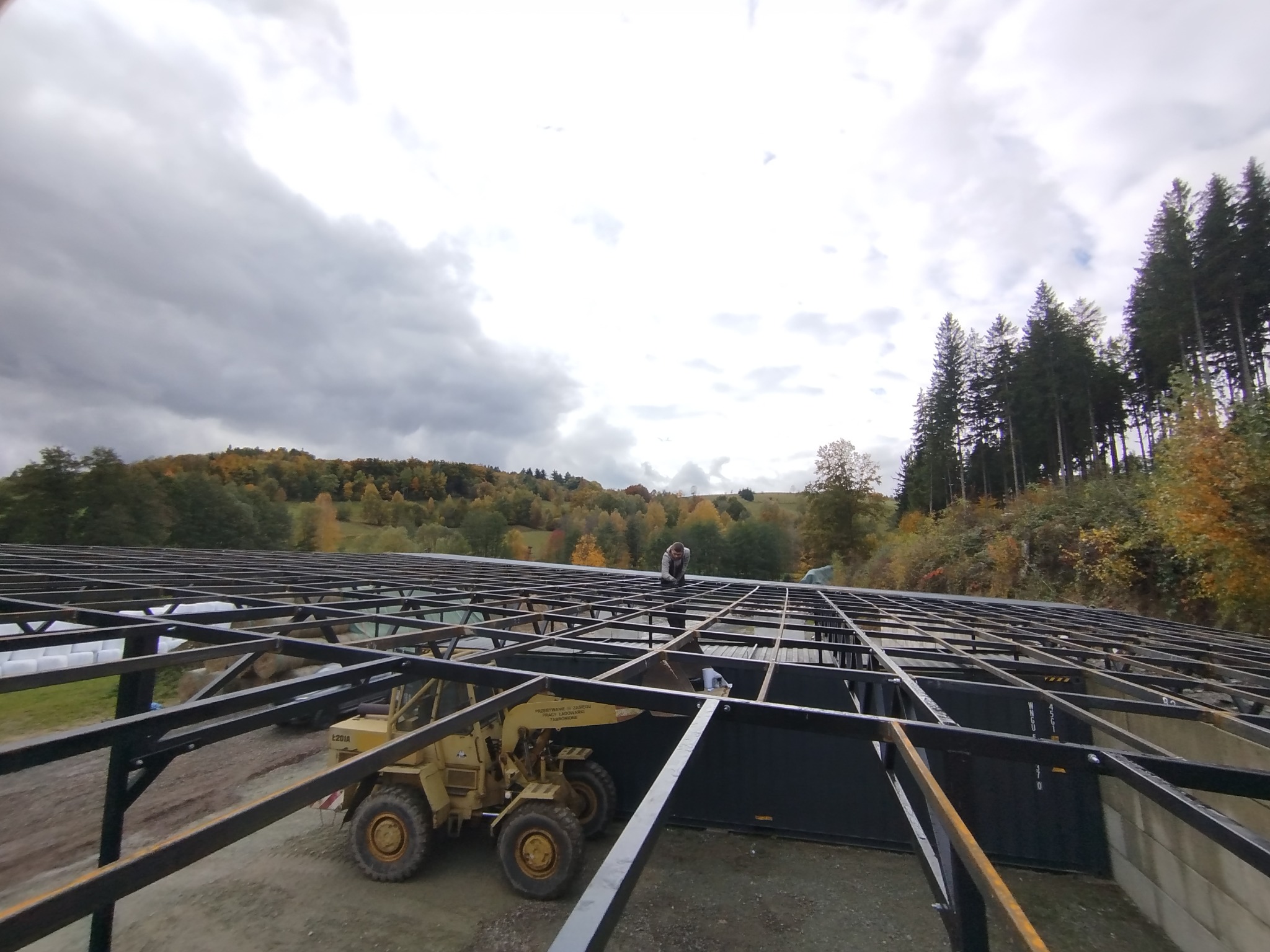 Konstrukcja stalowa hali w trakcie budowy, widoczny spawacz na dachu i żółty wózek widłowy. Tło stanowią drzewa i pochmurne niebo.