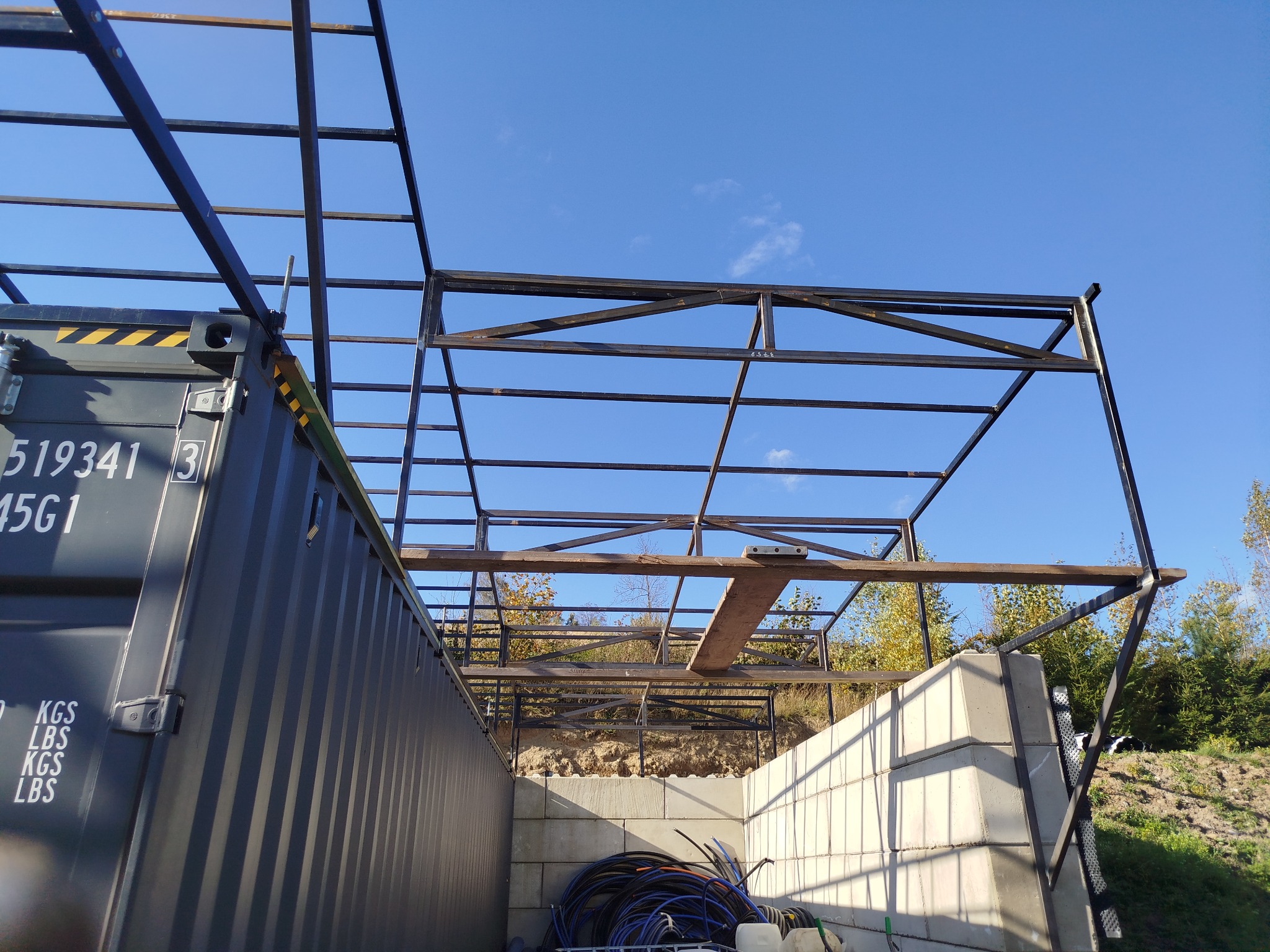 Konstrukcja stalowa w trakcie budowy hali, widoczna z dołu na tle błękitnego nieba. Elementy kontenera i betonowe bloczki w dolnej części kadru.