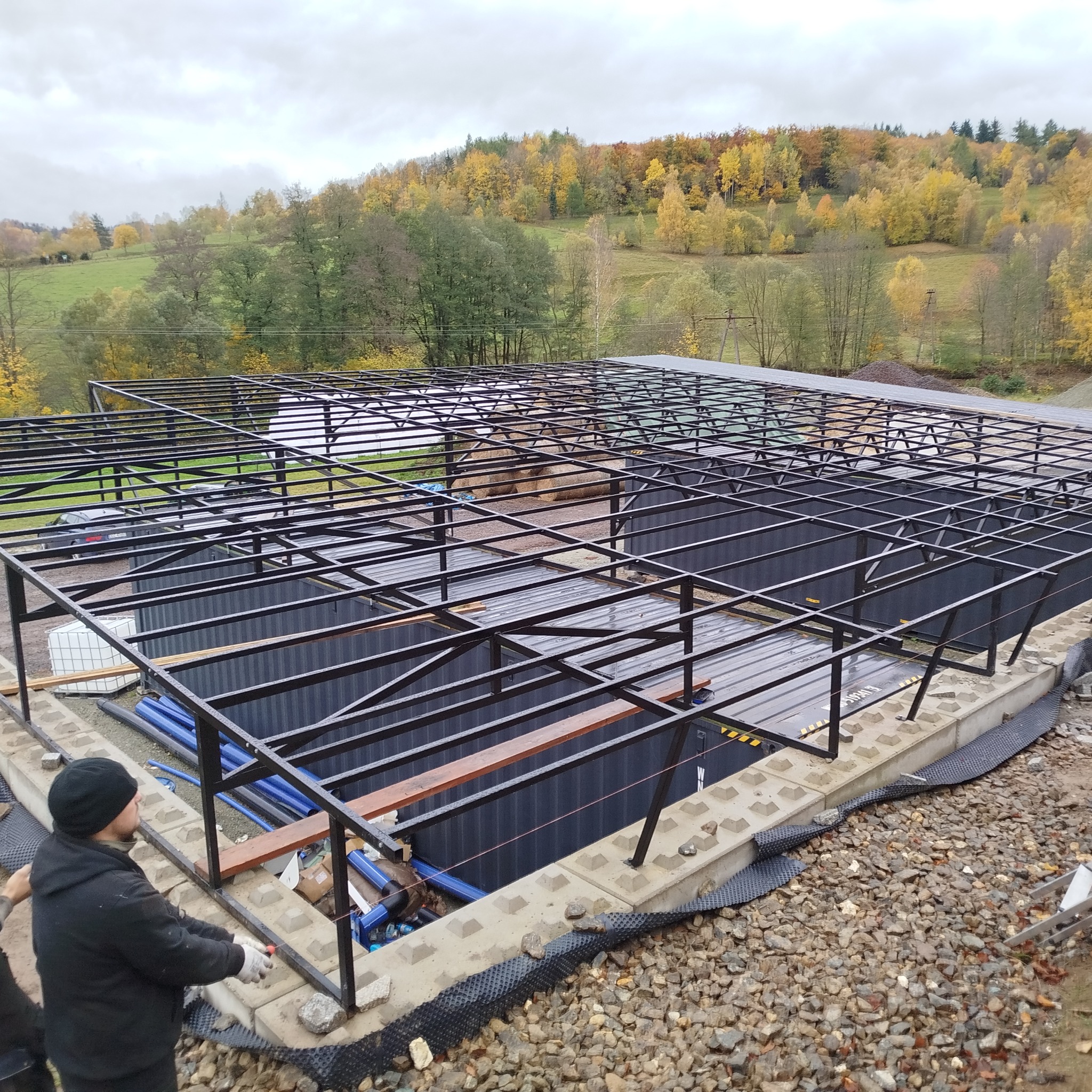 Stalowa konstrukcja hali w trakcie budowy, widok z góry na szkielet dachu i ściany z blachy trapezowej, pracownik w czapce, jesienny krajobraz w tle.