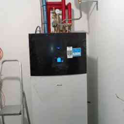 Zainstalowana pompa ciepła Daikin z widocznym zbiornikiem wyrównawczym, rurami w izolacji i drabinką aluminiową w rogu pomieszczenia.