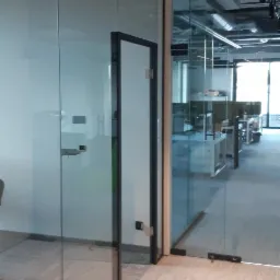 Szklane ścianki działowe z drzwiami w nowoczesnym biurze, widoczne metalowe okucia i jasna podłoga.