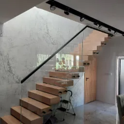 Nowoczesne schody drewniane z balustradą szklaną, wspornikowe, z czarnymi elementami mocującymi, oświetleniem punktowym i marmurową ścianą w tle, widoczne z dołu.