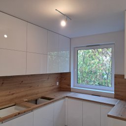 KJK Meble Krzysztof Krzysztofowicz - Jasna kuchnia z białymi szafkami i drewnianym blatem, widok na zieleń za oknem, oświetlenie halogenowe.