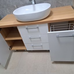 KJK Meble Krzysztof Krzysztofowicz - Biała umywalka nablatowa na szafce z drewnianym blatem, otwarte drzwiczki szafki po lewej stronie, szuflady z srebrnymi uchwytami, otwarta szafka z metalowym koszem w środku.