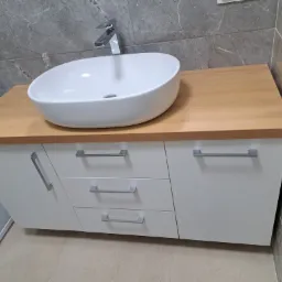 Łazienka z owalną umywalką nablatową, drewnianym blatem i białymi szafkami z szufladami oraz chromowanymi uchwytami, na tle szarych płytek imitujących marmur.