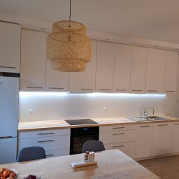 KJK Meble Krzysztof Krzysztofowicz - Nowoczesna kuchnia z białymi szafkami, drewnianym blatem, plecioną lampą i widocznym na pierwszym planie stołem z owocami.