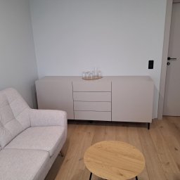 KJK Meble Krzysztof Krzysztofowicz - Jasnoszara komoda z trzema szufladami i szafkami, na której stoi zestaw szklanek, w minimalistycznym salonie z drewnianą podłogą i tapicerowaną sofą w odcieniach szarości.