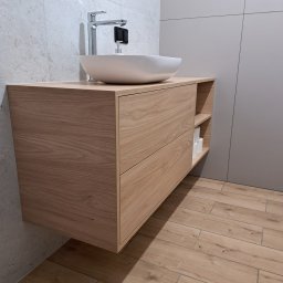 KJK Meble Krzysztof Krzysztofowicz - Drewniana szafka pod umywalkę z dwoma szufladami i otwartą półką boczną, na której widoczne są zwinięte ręczniki papierowe; umywalka nablatowa w kształcie misy i nowoczesna bateria.